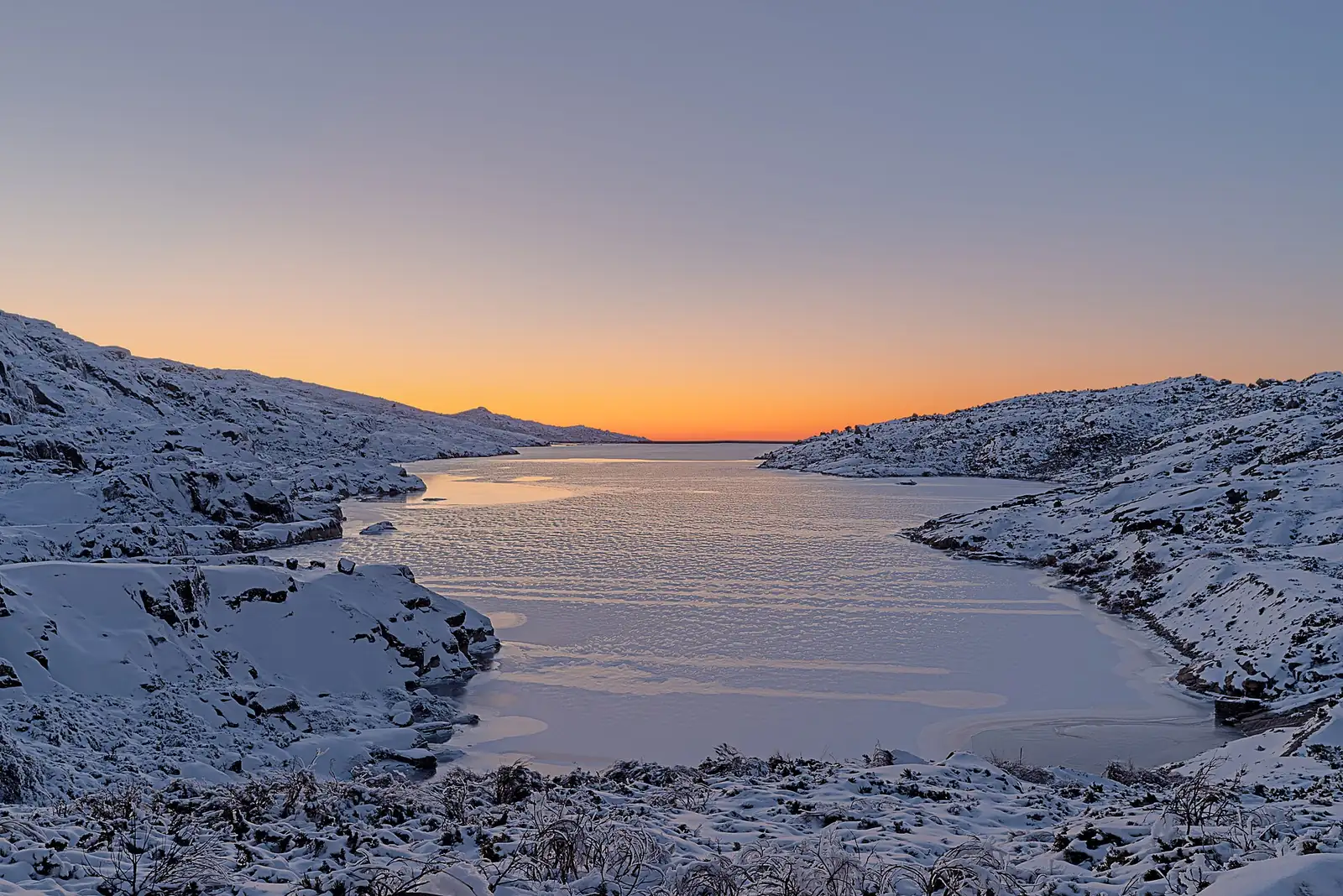 Lagoa Comprida gelada na Serra da Estrela, com neve nas margens e o céu laranja ao fim do dia