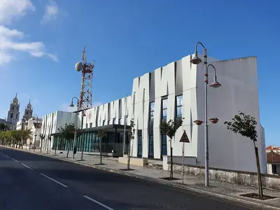 Edifício branco da Loja do Cidadão de Mafra, com as torres do Palácio Nacional de Mafra ao fundo