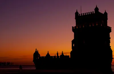A Torre de Belém em silhueta contra o céu laranja e roxo do pôr do sol, em Lisboa